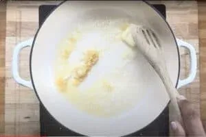 Person stirring butter and garlic in a white pot with wooden spoon on stovetop.